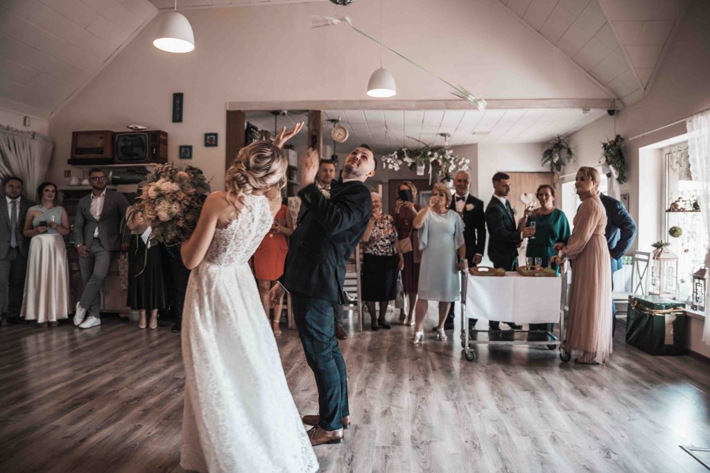 Para młoda wśród gości podczas wesela, para młoda rzuca kieiliszkami, a goście obserwują moment z uśmiechem.