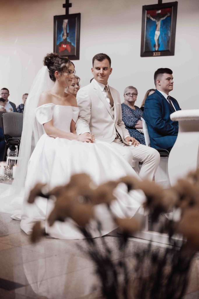 Para młoda siedzi obok siebie w kościele podczas ceremonii ślubnej, trzymają się za ręce, w tle zgromadzeni goście.
